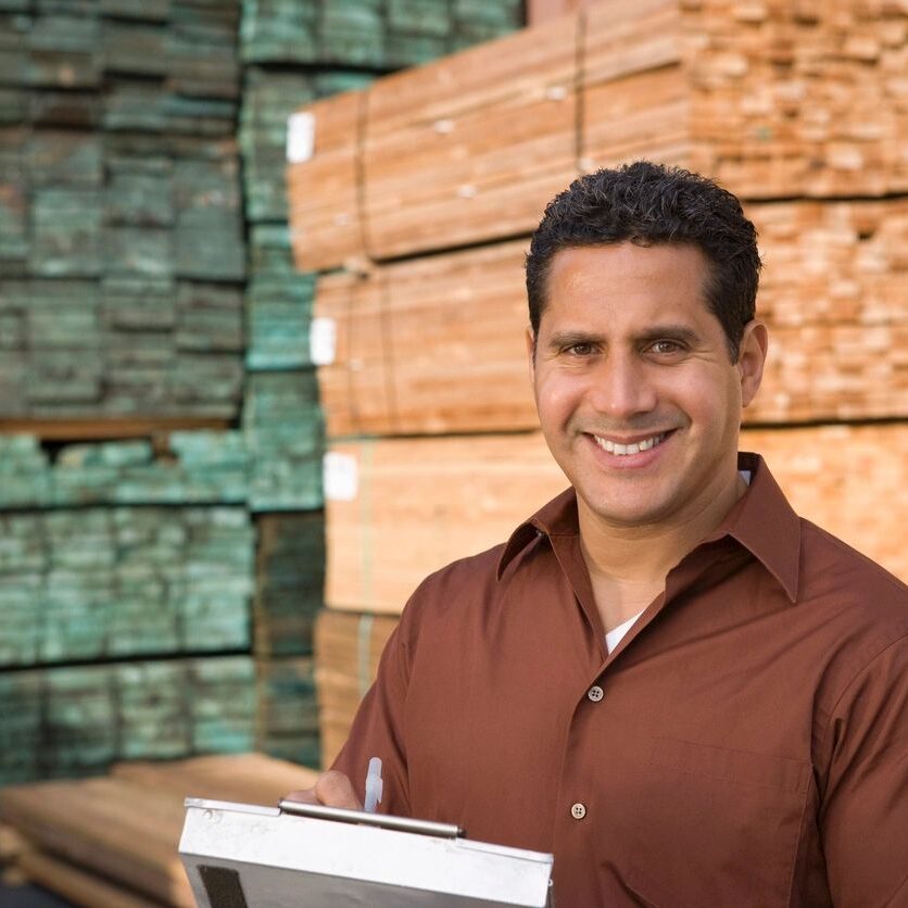 Smiling man holding a clipboard in a lumber yard.
